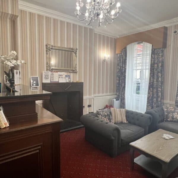 A cozy lounge with patterned wallpaper, a chandelier, tufted gray sofas, a wooden coffee table, red carpet, a fireplace with a mirror above, and a window with sheer curtains and a brown valance.