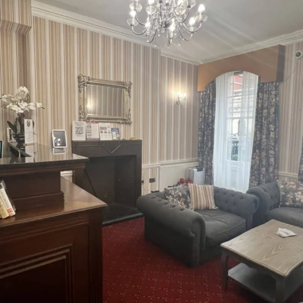A cozy lounge with patterned wallpaper, a chandelier, tufted gray sofas, a wooden coffee table, red carpet, a fireplace with a mirror above, and a window with sheer curtains and a brown valance.