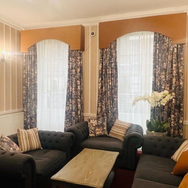 A cozy living room with striped wallpaper, two windows with floral curtains, two dark tufted sofas with patterned cushions, a wooden coffee table, and a white orchid plant in the corner.