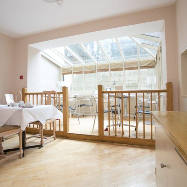 Bright dining area with wooden chairs and tables, leading to a sunlit conservatory with more seating. A counter with cereal boxes is visible on the right, and the room has pale walls and wooden flooring.
