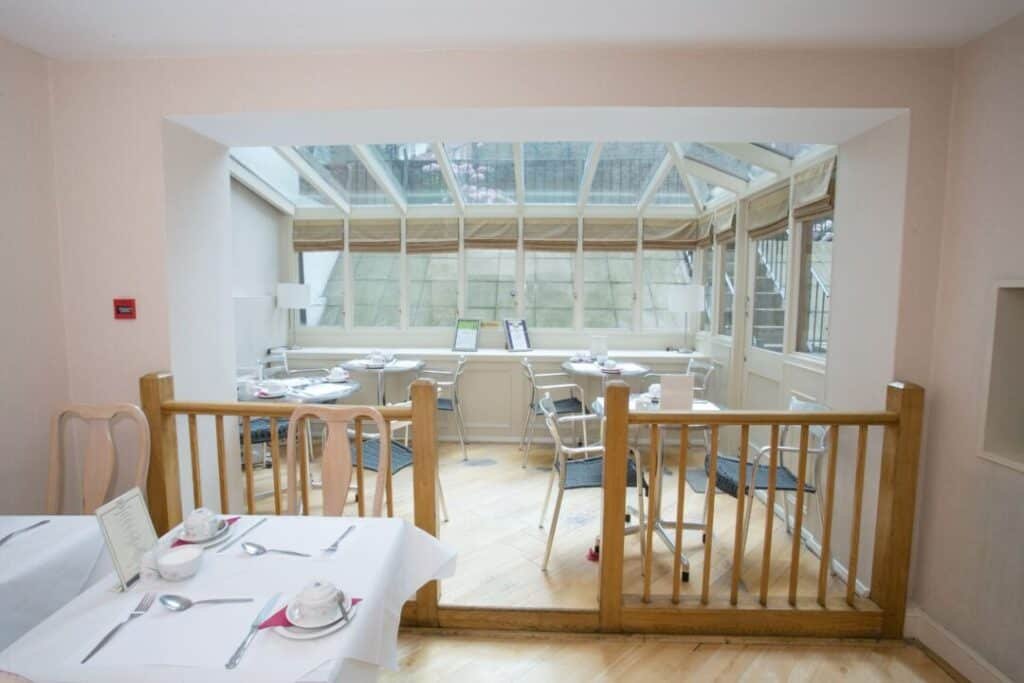 A bright dining area with white tablecloths, chairs, and place settings, separated by a wooden railing. The room extends into a glass-ceilinged sunroom filled with more tables and chairs, creating an airy atmosphere.