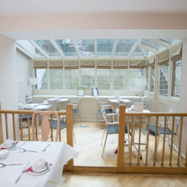 A bright dining area with white tablecloths, chairs, and place settings, separated by a wooden railing. The room extends into a glass-ceilinged sunroom filled with more tables and chairs, creating an airy atmosphere.
