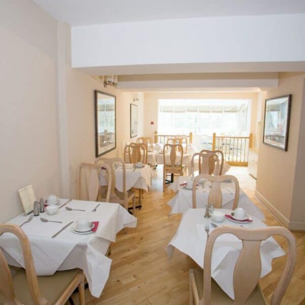 A small, bright dining area with wooden chairs and tables covered in white tablecloths, set with cups, saucers, and cutlery. Large windows let in natural light at the far end of the room.