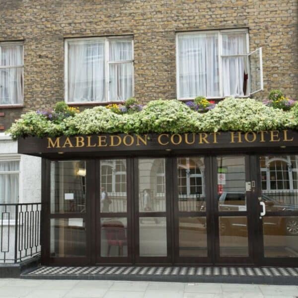 Entrance of Mabledon Court Hotel with glass doors, a dark awning featuring the hotel name in gold letters, and a row of flowers above. Brick building with windows and white curtains in the background.