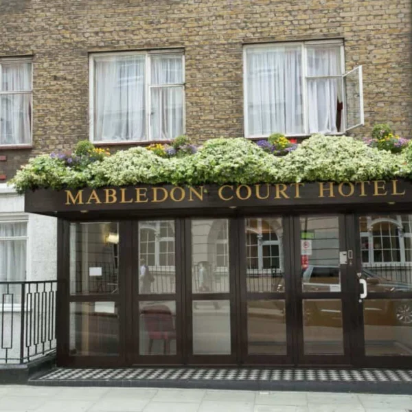 Entrance of Mabledon Court Hotel with glass doors, a dark awning featuring the hotel name in gold letters, and a row of flowers above. Brick building with windows and white curtains in the background.