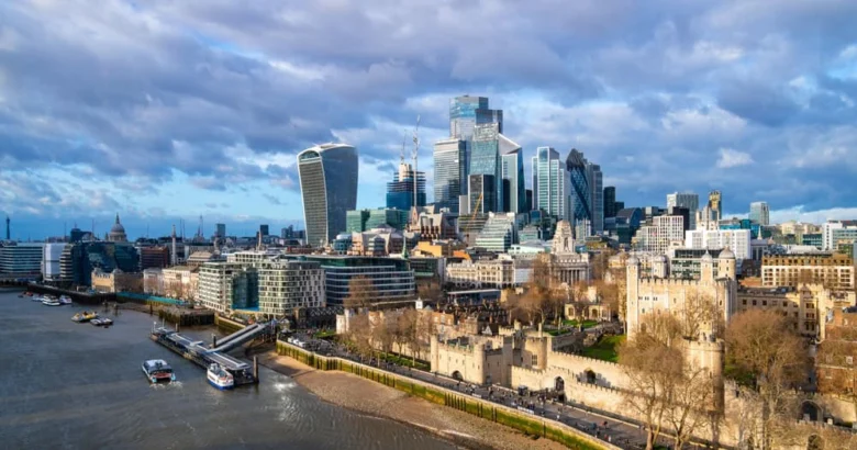 A view of London’s skyline with modern skyscrapers, historic buildings, the Tower of London in the foreground, and the River Thames on the left under a partly cloudy sky.