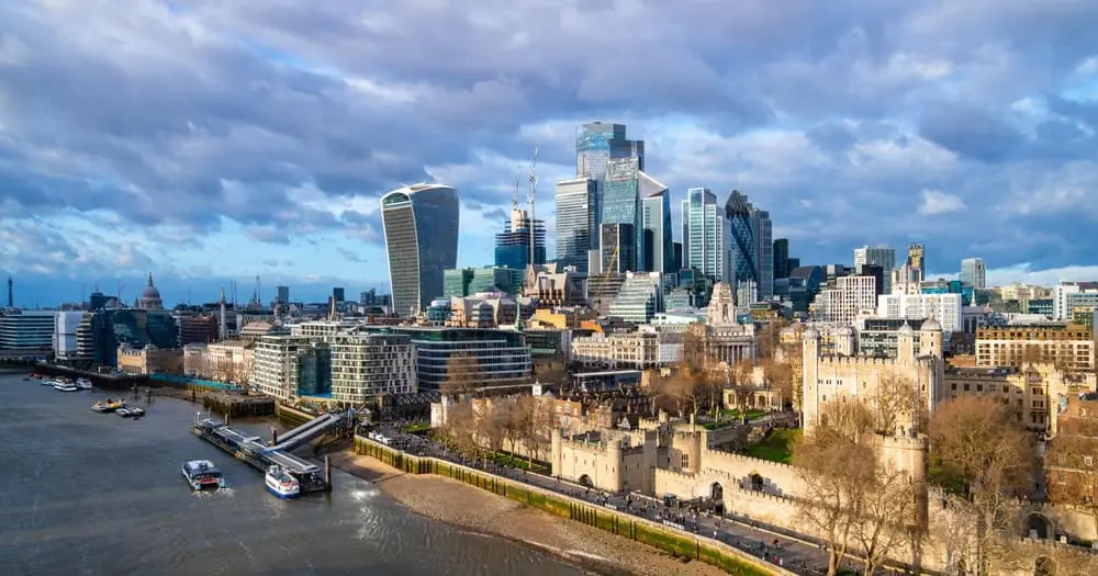 A view of London’s skyline with modern skyscrapers, historic buildings, the Tower of London in the foreground, and the River Thames on the left under a partly cloudy sky.