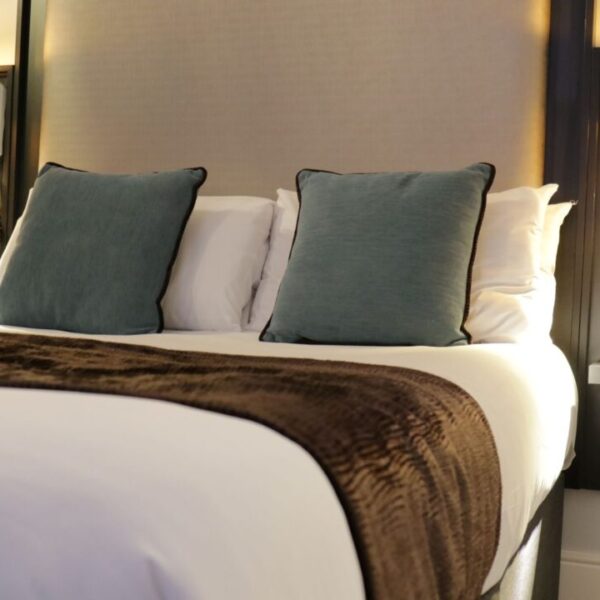 A neatly made hotel bed with white linens, a brown throw blanket, and two blue accent pillows. Wall-mounted lamps and built-in electrical outlets are on either side of the headboard.