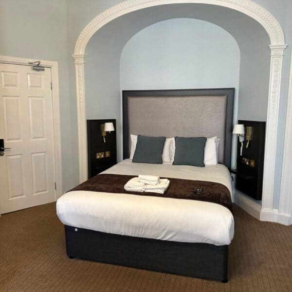 A neatly made double bed with white linens, a brown throw, and teal cushions is set in an arched alcove with wall lights, flanked by two doors in a tidy, modern bedroom with beige carpet. Towels and a TV remote rest on the bed.