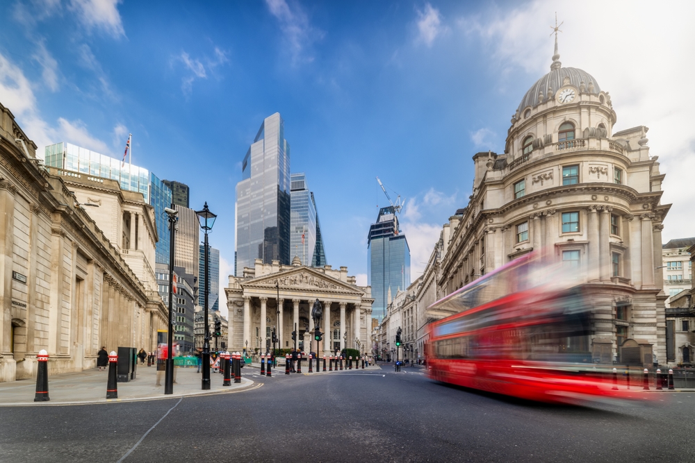 A red double-decker bus speeds past historic and modern buildings, including the Royal Exchange, in the financial district of London under a blue sky with scattered clouds.