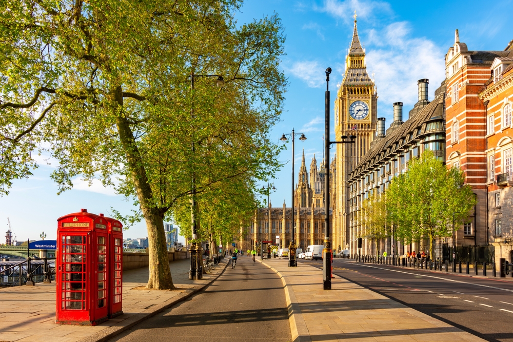 A sunny street scene in London featuring a red telephone booth, trees lining the sidewalk, and the iconic Big Ben clock tower and Houses of Parliament in the background under a blue sky.