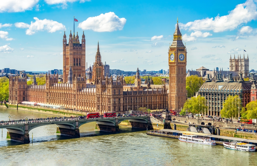 The Palace of Westminster and Big Ben beside the River Thames in London on a sunny day, with red double-decker buses crossing Westminster Bridge and boats on the water.