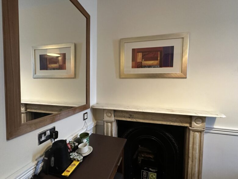 A desk with a kettle, cups, and tea supplies sits beside a white marble fireplace. A large mirror and a framed abstract artwork hang on the wall above and beside the desk.