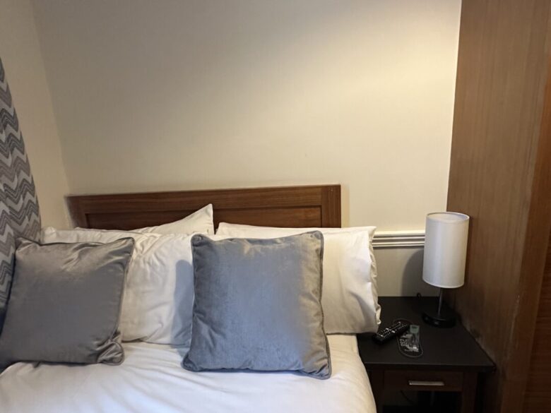 A neatly made bed with white sheets and two gray pillows is next to a wooden nightstand holding a lamp, remote controls, and a pair of glasses. The wall behind is plain and light colored.