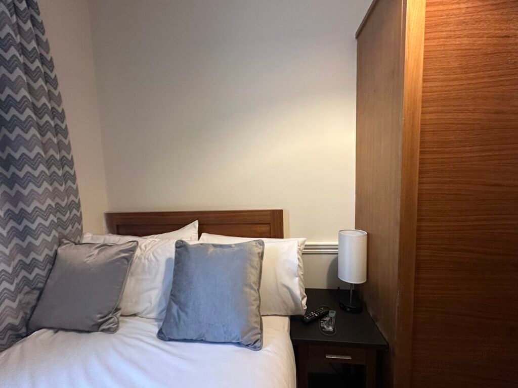 A neatly made bed with white sheets and three gray pillows next to a patterned curtain. A wooden nightstand holds a white lamp, phone, and glasses, beside a tall wooden wardrobe.