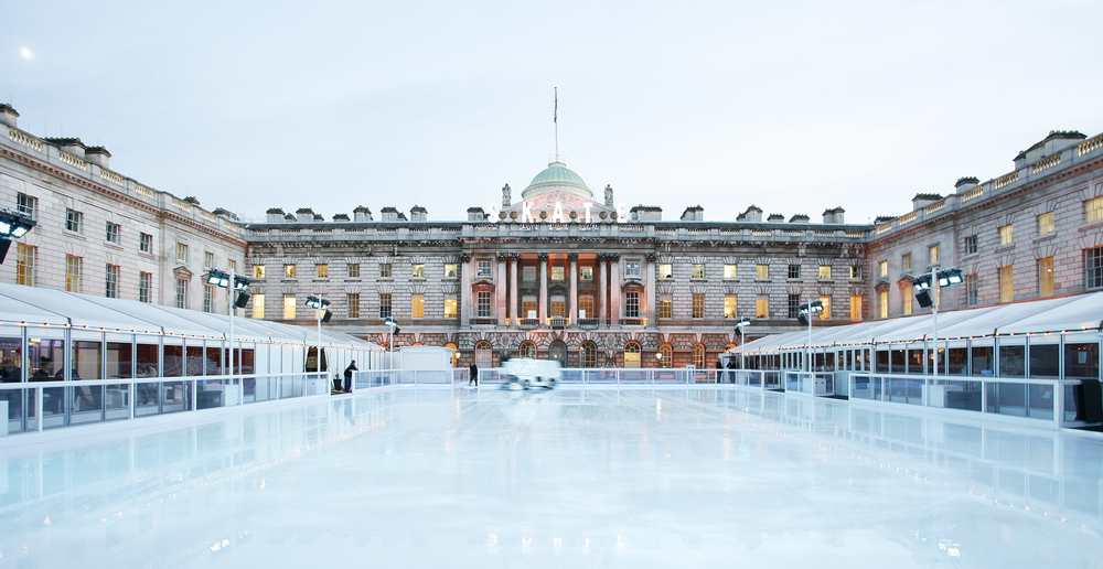A large, empty outdoor ice skating rink is set in front of a grand historic building with columns and a dome, surrounded by covered walkways and illuminated by soft lights.