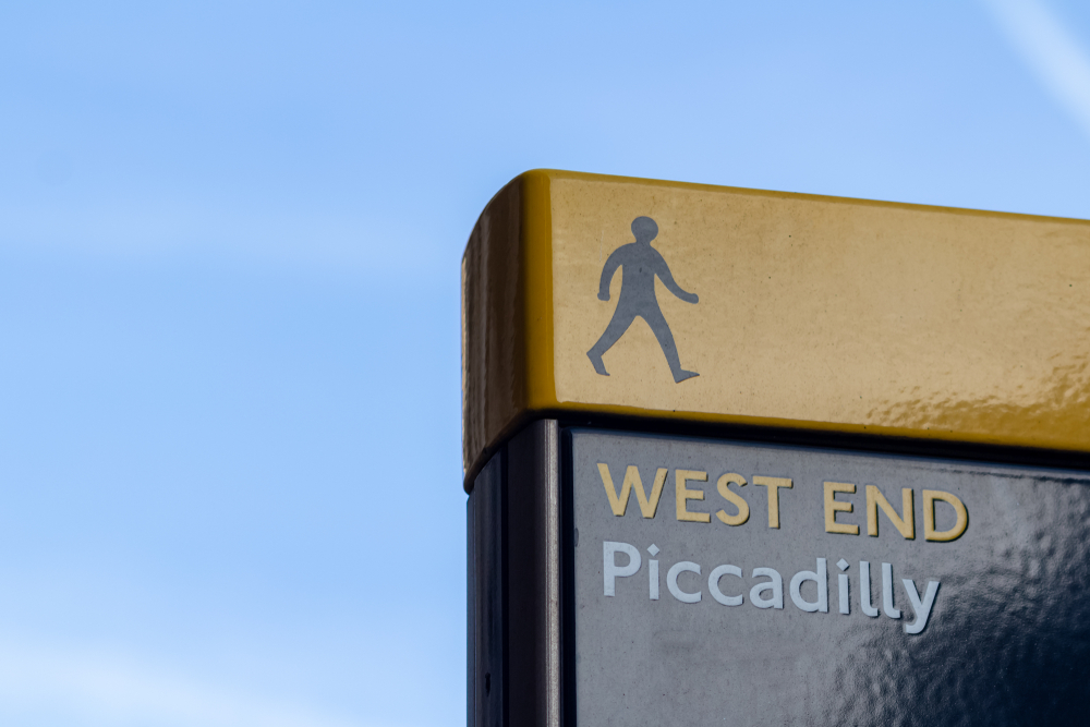 A sign against a blue sky reads "WEST END Piccadilly" with a walking person symbol above the text, indicating a pedestrian direction or area.