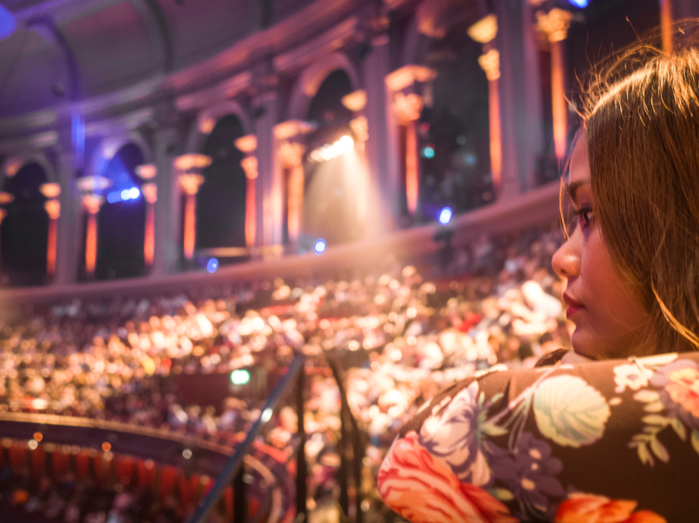 A woman with long hair and a floral top looks toward a brightly lit auditorium filled with seated people, arched windows, and glowing lights in the background.