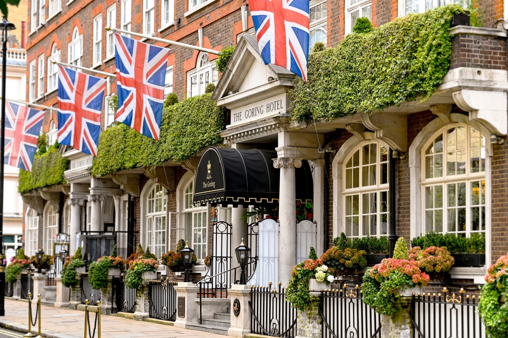The exterior of The Goring Hotel, adorned with British flags, lush greenery, and potted flowers along the entrance. The building features large arched windows and a black canopy over the main door.