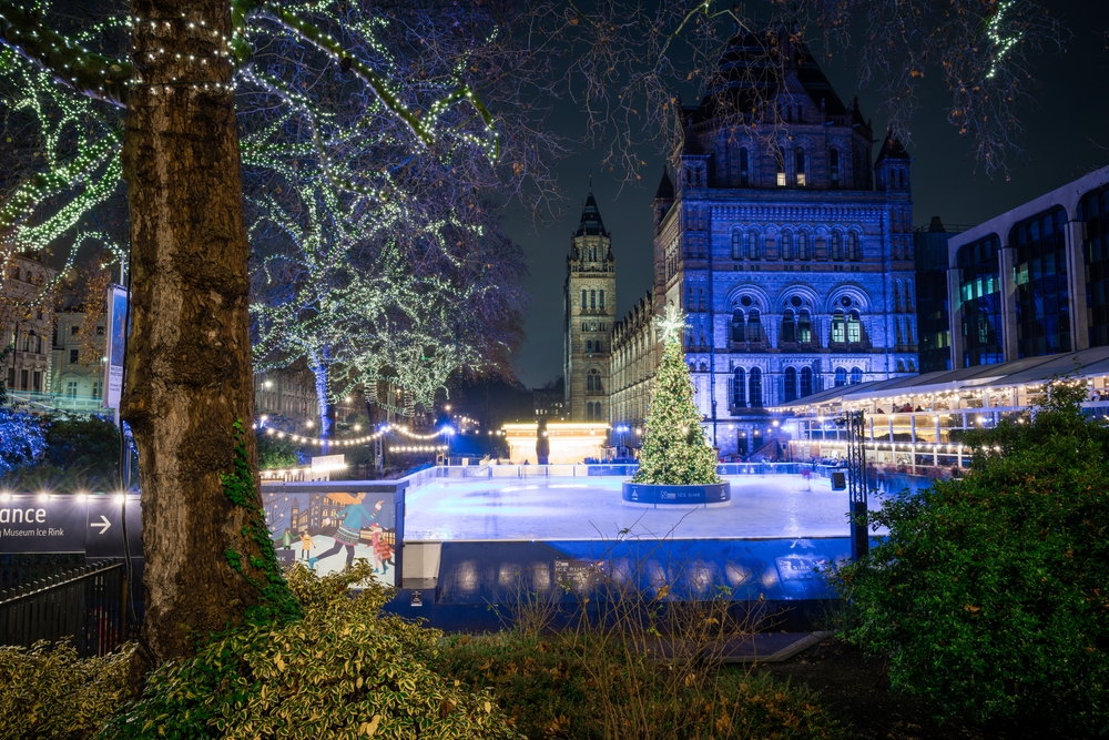 A festive outdoor ice skating rink lit with string lights and a large Christmas tree in front of the illuminated Natural History Museum in London at night. Trees and bushes frame the scene.