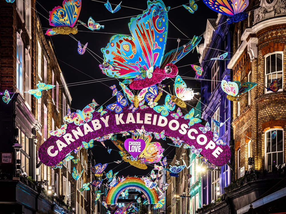 A vibrant street scene at night features colorful butterfly decorations, a rainbow, and a glittery sign reading “Carnaby Kaleidoscope” with “Choose Love” below, illuminating a lively urban setting with festive lights.