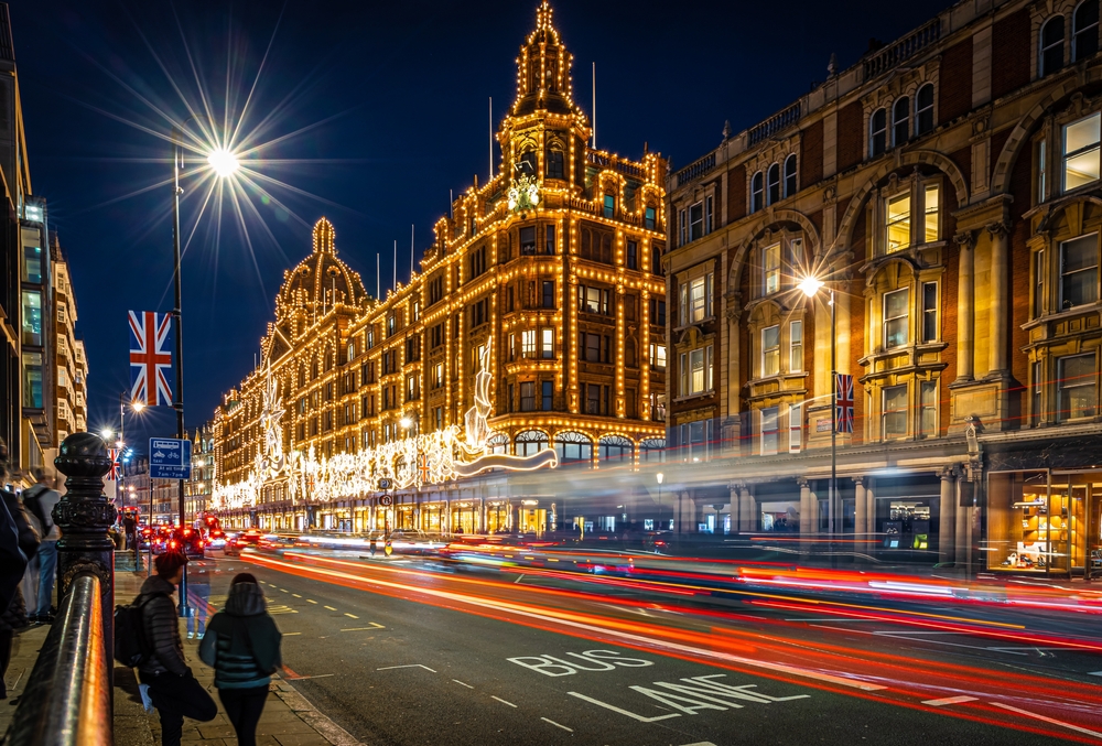 A busy city street at night with cars’ light trails passing by an ornate, brightly illuminated building decorated with festive lights; people walk along the sidewalk and Union Jack flags hang on lamp posts.