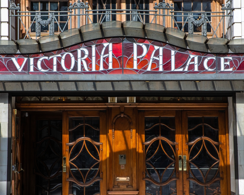 Entrance to the Victoria Palace, featuring a stained glass sign with the venue's name above intricately designed wooden doors.