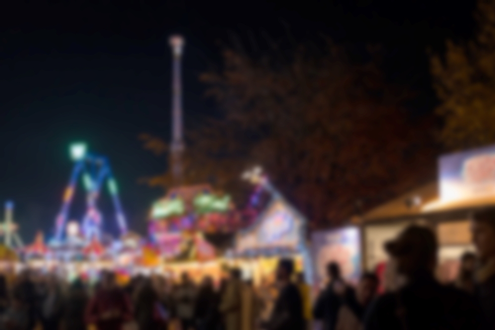 A blurry nighttime photo of a busy fairground with colorful lights, rides, and crowds of people. The scene is lively but out of focus, emphasizing the vibrant atmosphere rather than details.