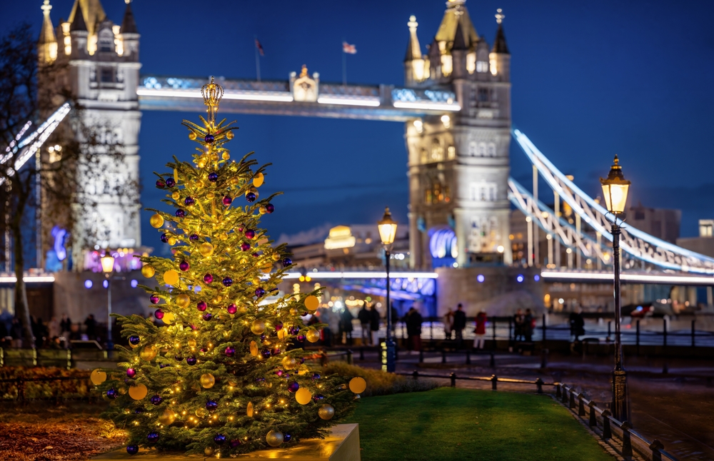 A decorated Christmas tree with yellow and purple ornaments glows in front of the illuminated Tower Bridge in London at night, with people walking and street lamps lit.