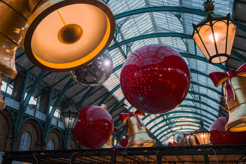 Large red Christmas ornaments, gold bells, and festive decorations hang from the ceiling of a glass-roofed indoor market or atrium, illuminated by warm light from hanging chandeliers.