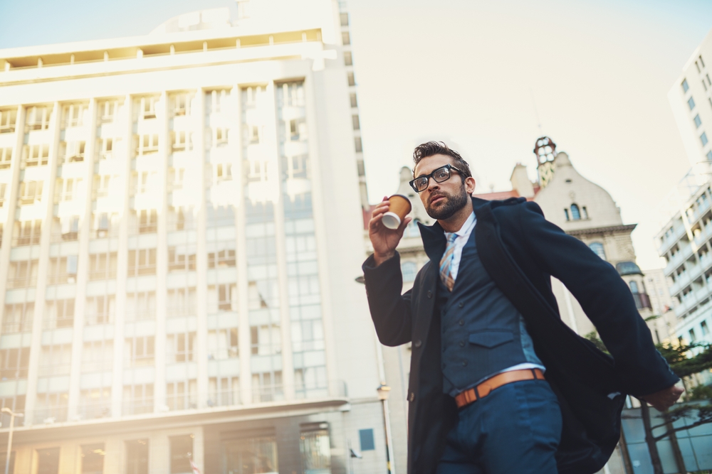 A man in a suit and overcoat walks confidently outdoors in a city, holding a coffee cup. Tall, sunlit buildings are in the background, and he is wearing glasses and looking slightly upward.
