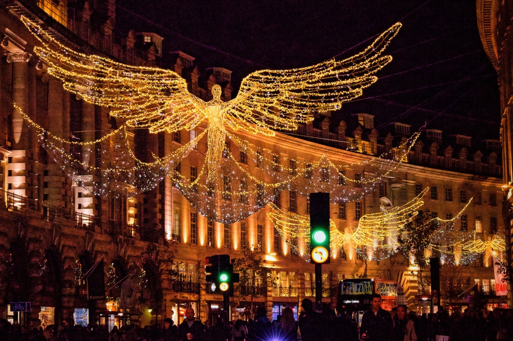 Large angel-shaped holiday lights are suspended above a busy city street at night, with historic buildings in the background and people walking below. The scene is festive and vibrant, with green traffic lights visible.