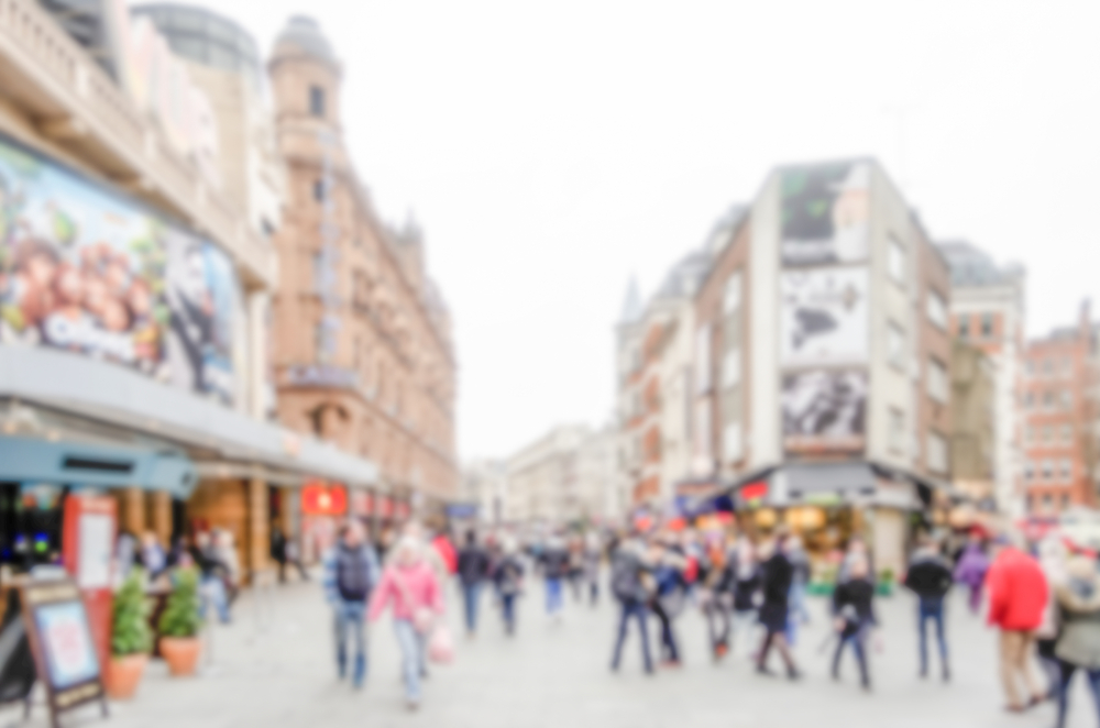 A busy city street with many people walking and historic buildings in the background; the image is blurred, obscuring faces and details.