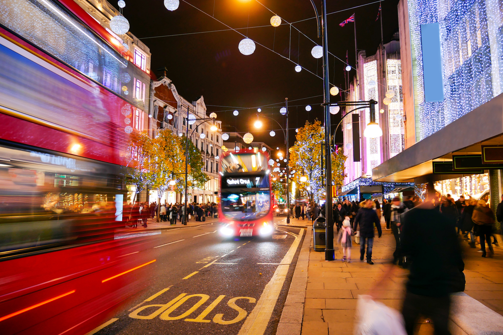 A busy city street at night with two red double-decker buses in motion, festive lights hanging above, illuminated shops, and crowds of people walking along the sidewalk. The word “STOP” is painted on the road.
