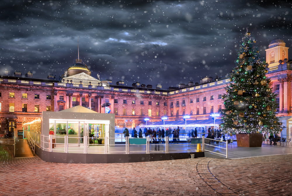 A festive ice skating rink with people skating, a large decorated Christmas tree, and a historic building illuminated by colorful lights at night; snow is gently falling.