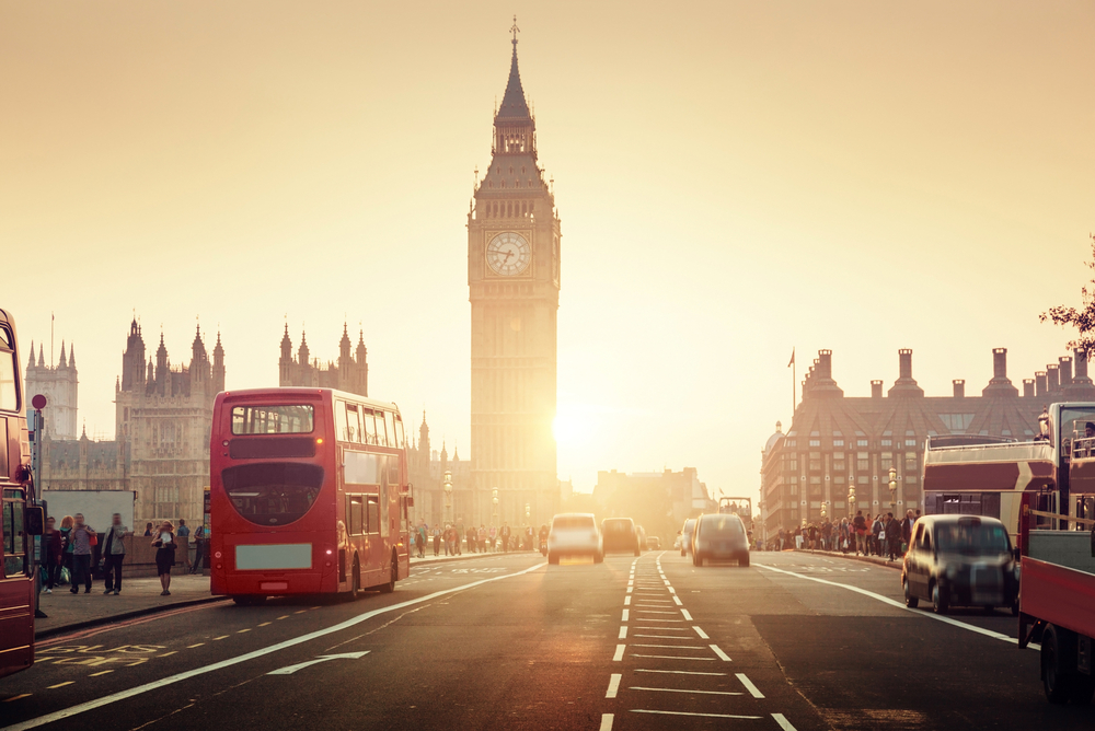 A busy London street at sunset with red double-decker buses, cars, and people. The iconic Big Ben clock tower and the Houses of Parliament are prominent in the background.