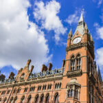 A historic red brick building with ornate architecture and a tall clock tower under a bright blue sky with fluffy white clouds.