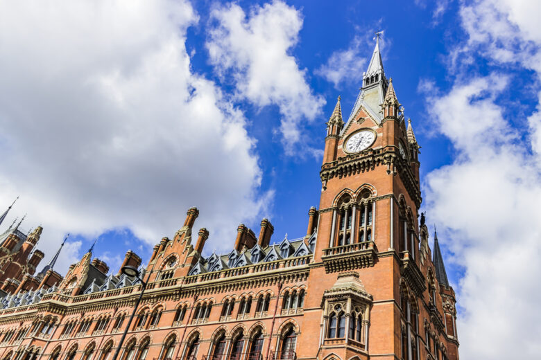 A historic red brick building with ornate architecture and a tall clock tower under a bright blue sky with fluffy white clouds.