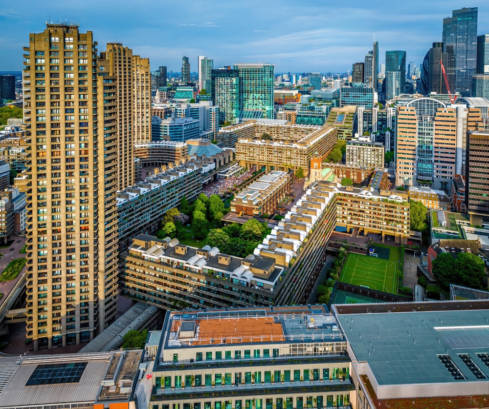 Aerial view of a cityscape with tall modern buildings, residential complexes, and green spaces, including a sports field, under a blue sky in London’s Barbican area.