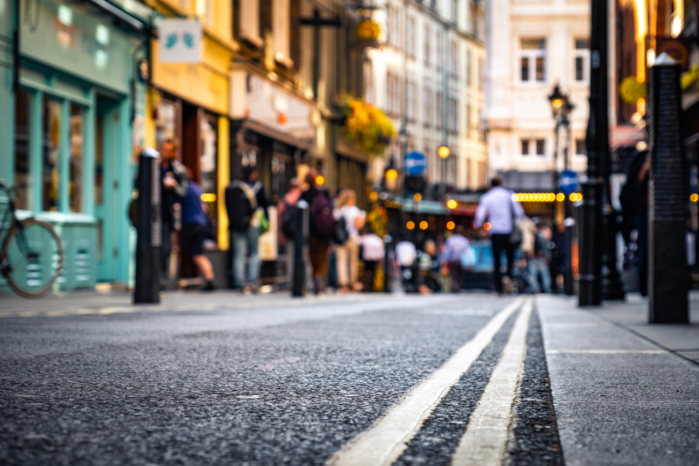 Street-level view of a city street with yellow lines, busy with people walking and standing outside colorful shops and cafes; the background is blurred, giving a vibrant, lively urban atmosphere.