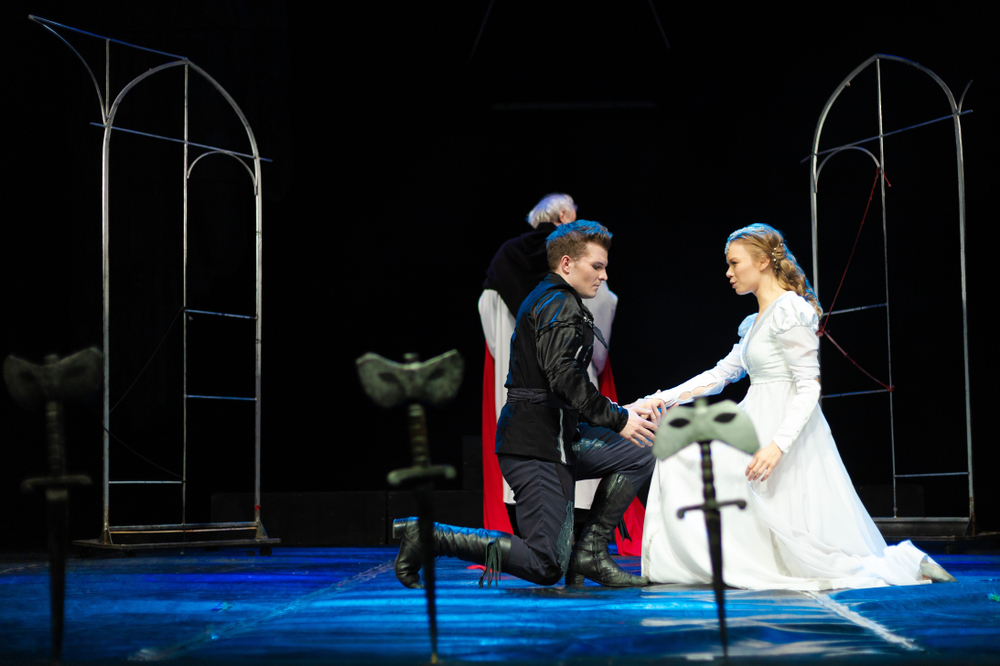 A man in a black costume kneels and holds hands with a woman in a long white dress on a theater stage. Two arch frames and sword props are in the foreground; another person stands in the background, partially turned away.