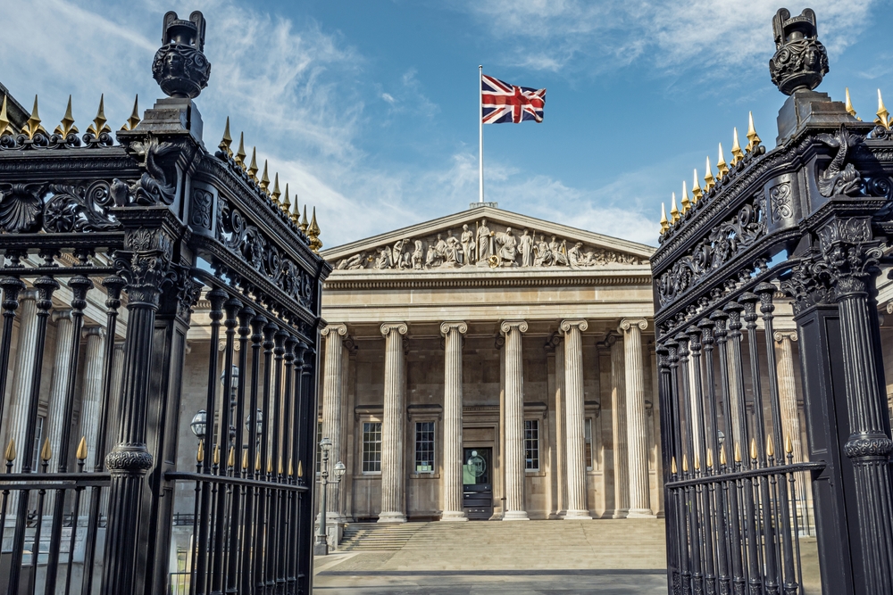 A grand neoclassical building with tall columns and a British flag on top is seen through ornate black iron gates under a blue sky with light clouds.