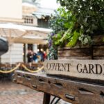 Close-up of a wooden sign reading "Covent Garden" with green plants behind it; in the background, there are tables, chairs, and people in an outdoor plaza setting.