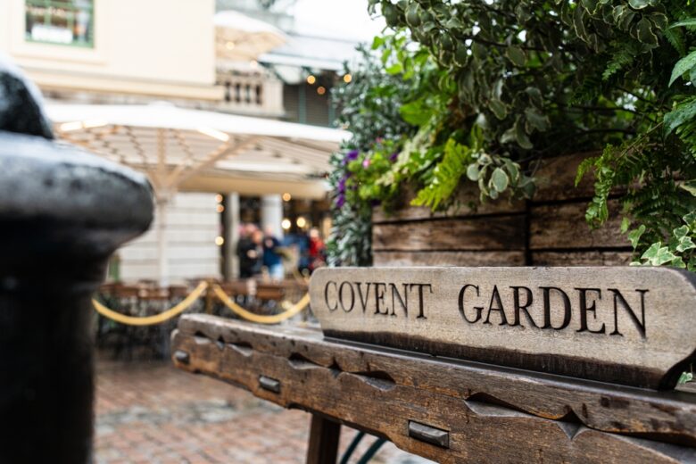 Close-up of a wooden sign reading "Covent Garden" with green plants behind it; in the background, there are tables, chairs, and people in an outdoor plaza setting.