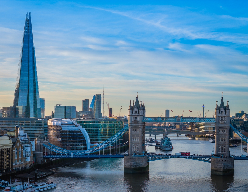 A view of London’s Tower Bridge spanning the River Thames, with the modern glass Shard skyscraper and other city buildings visible under a blue sky.