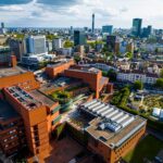 Aerial view of the British Library’s distinctive red-brick buildings, surrounded by city streets, greenery, and modern high-rise buildings in central London under a partly cloudy sky.
