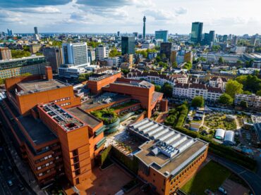 Aerial view of the British Library’s distinctive red-brick buildings, surrounded by city streets, greenery, and modern high-rise buildings in central London under a partly cloudy sky.
