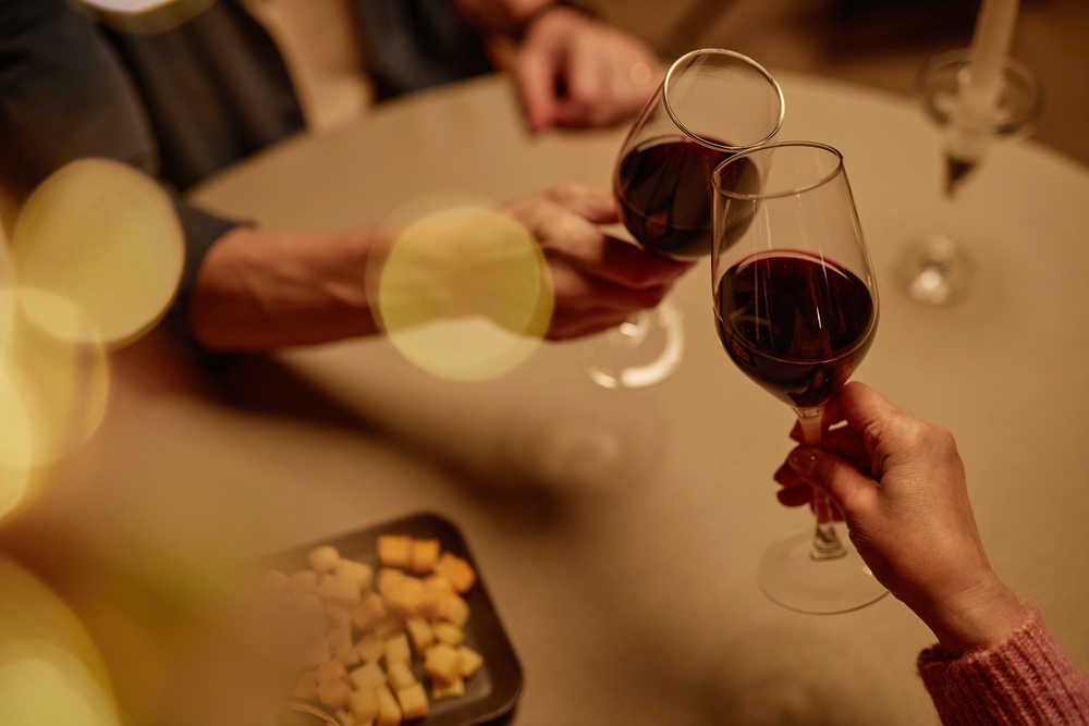 Two people clinking glasses of red wine over a round table, with a plate of cheese and blurred warm lights creating a cozy atmosphere.