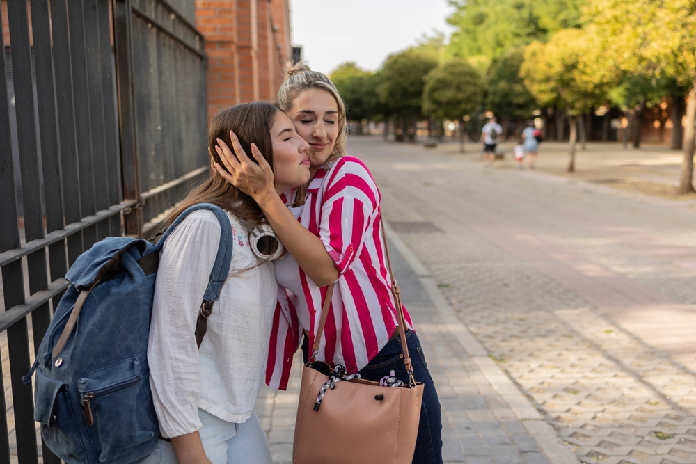 A woman in a striped shirt kisses a young girl with a backpack on the cheek as they hug on a sidewalk lined with trees and brick buildings.