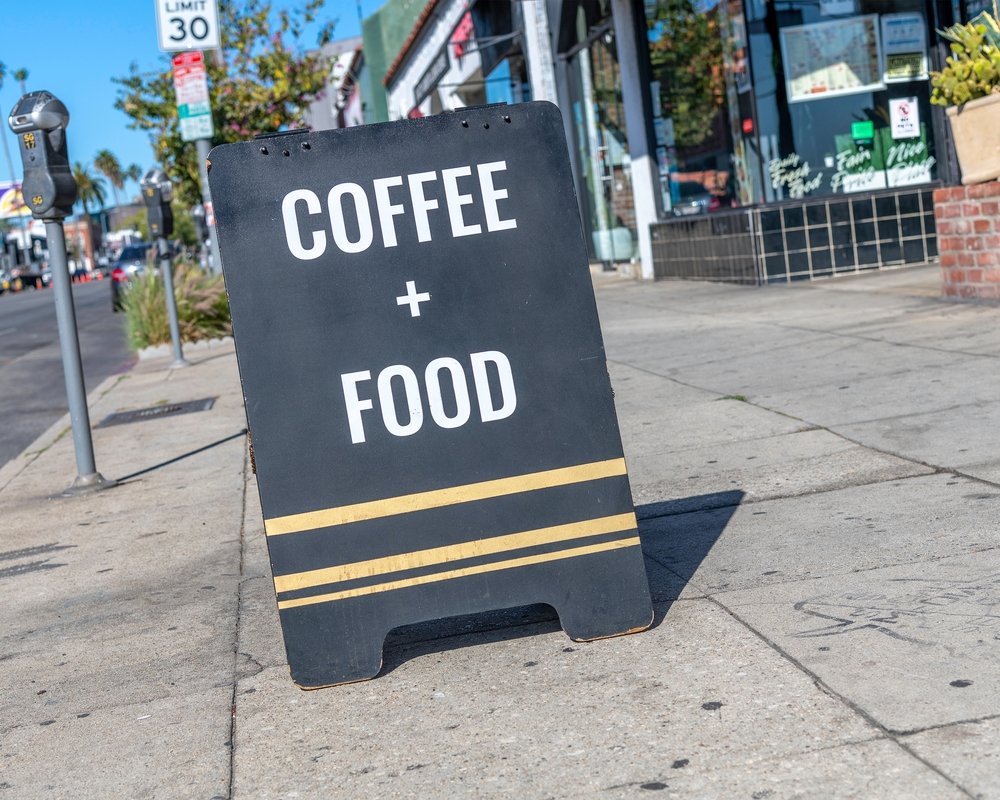 A sidewalk sign reads "COFFEE + FOOD" in bold white letters, placed on a city street near shops and a parked car. The sign is black with two yellow stripes near the bottom.
